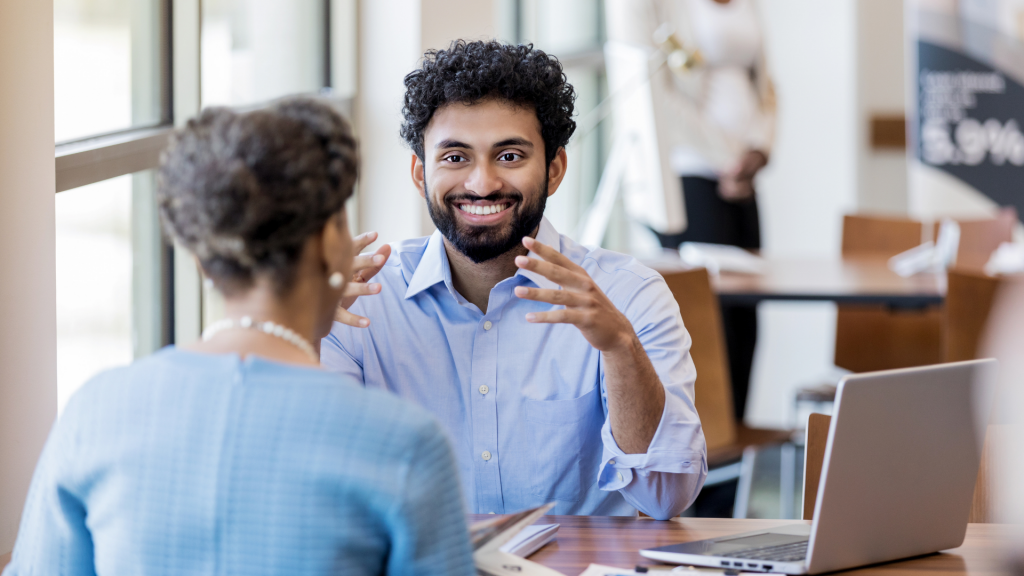 Male person of color banker sits across the desk from a woman person of color customer while talking