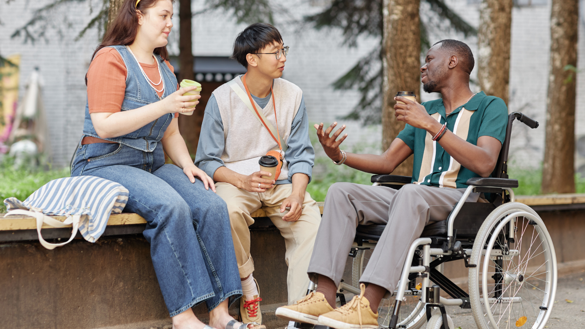 Group of diverse young friends, including a white woman, Asian man, and Black man in a wheel chair sit ourdoors and talk
